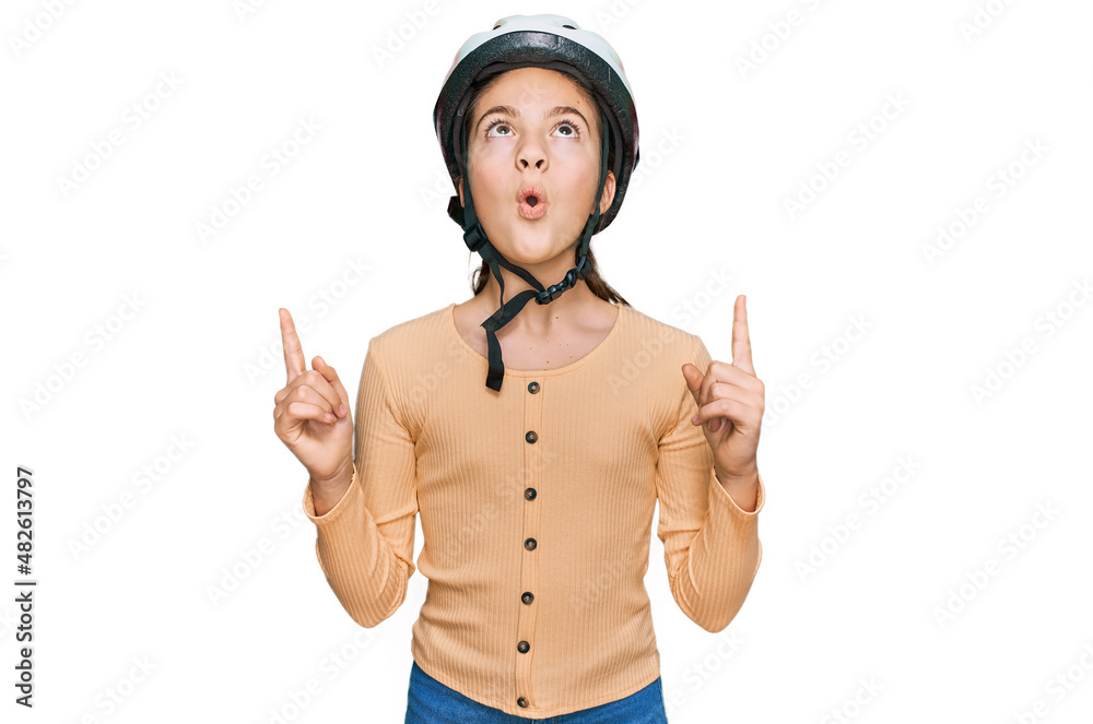 Beautiful brunette little girl wearing bike helmet amazed and surprised looking up and pointing with fingers and raised arms. Beautiful brunette little girl wearing bike helmet amazed and surprised looking up and pointing with fingers and raised arms.