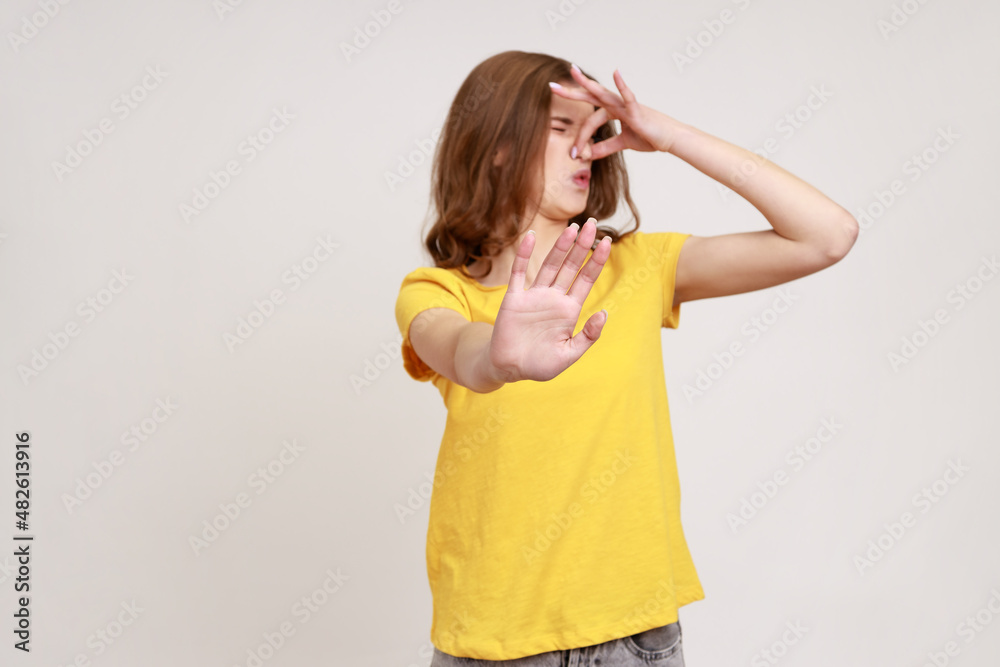 Portrait of displeased teenager girl in yellow T-shirt grimacing with disgust, pinching nose with fingers to avoid bad smell and showing stop gesture. Indoor studio shot isolated on gray background. Portrait of displeased teenager girl in yellow T-shirt grimacing with disgust, pinching nose with fingers to avoid bad smell and showing stop gesture. Indoor studio shot isolated on gray background.