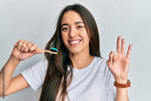 Fototapeta Naklejka Na Ścianę i Meble -  Young hispanic girl holding toothbrush with toothpaste doing ok sign with fingers, smiling friendly gesturing excellent symbol