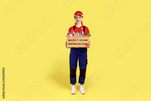Full length portrait of positive satisfied woman volunteer standing with box full of empty plastic bottles, wearing overalls and red cap. Indoor studio shot isolated on yellow background.