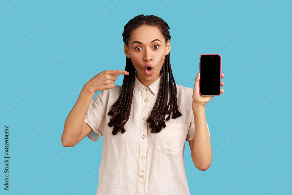 Surprised beautiful woman with dreadlocks having scared expression, points at mobile phone, demonstrates empty screen of gadget, wearing white shirt. Indoor studio shot isolated on blue background. Surprised beautiful woman with dreadlocks having scared expression, points at mobile phone, demonstrates empty screen of gadget, wearing white shirt. Indoor studio shot isolated on blue background.