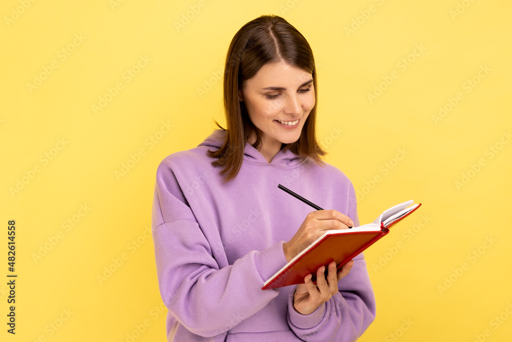 Portrait of happy inspired woman smiling and writing down interesting idea in notebook, making plans, to-do list, wearing purple hoodie. Indoor studio shot isolated on yellow background. Portrait of happy inspired woman smiling and writing down interesting idea in notebook, making plans, to-do list, wearing purple hoodie. Indoor studio shot isolated on yellow background.