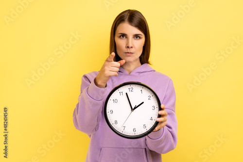 Strict dark haired young adult woman holding wall clock in hands and pointing to camera at you, time management, you are late, wearing purple hoodie. Indoor studio shot isolated on yellow background.