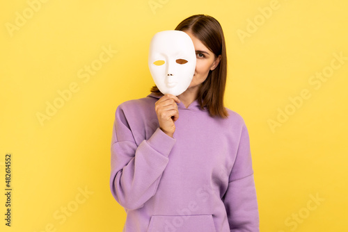Fotografija Woman with dark hair removing white mask from face showing his smiling expression, good mood, pretending to be another person, wearing purple hoodie