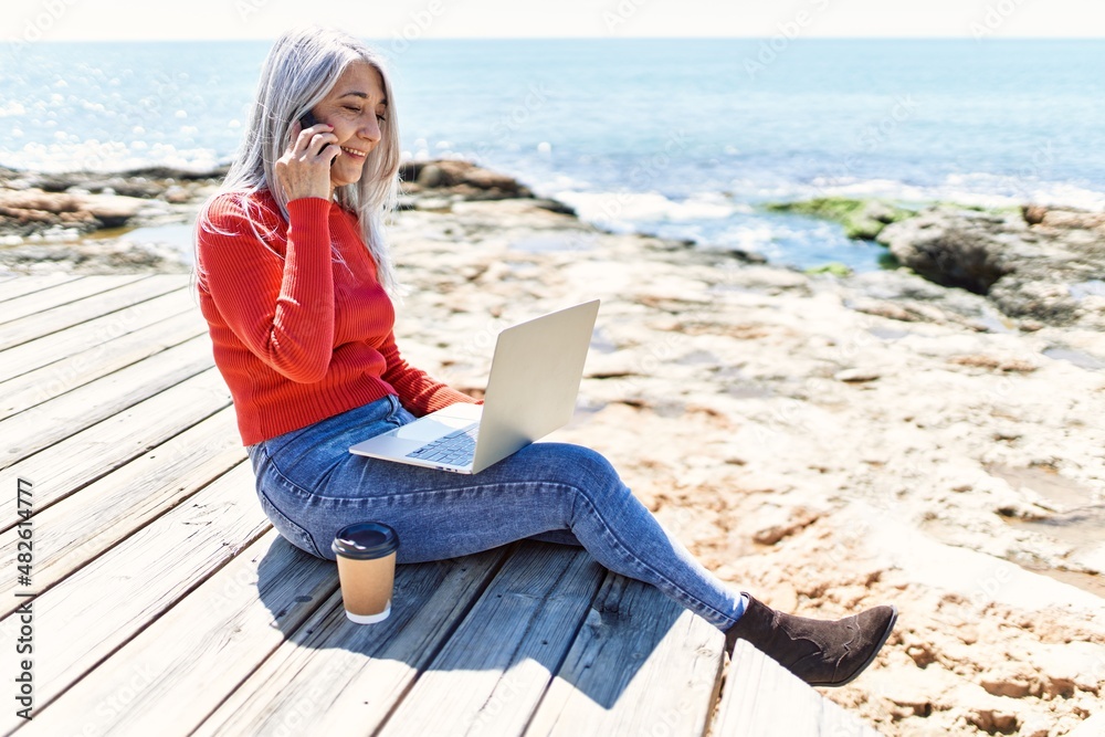 Middle age grey-haired woman using laptop and talking on the smartphone at the beach.