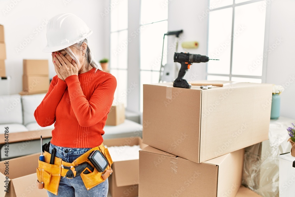 Middle age grey-haired woman wearing hardhat standing at new home with sad expression covering face with hands while crying. depression concept. Middle age grey-haired woman wearing hardhat standing at new home with sad expression covering face with hands while crying. depression concept.