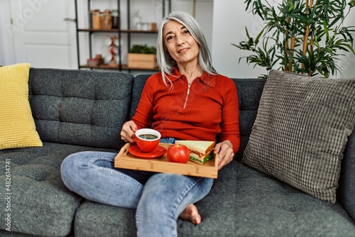 Middle age grey-haired woman having breakfast sitting on sofa at home photo