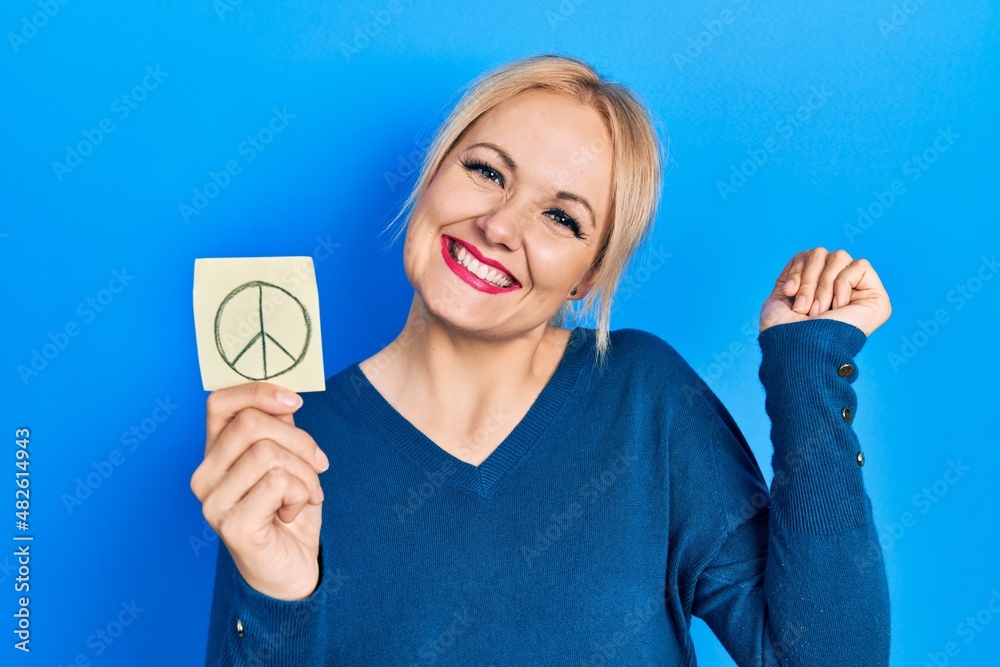 Young blonde woman holding peace symbol reminder paper screaming proud, celebrating victory and success very excited with raised arm Young blonde woman holding peace symbol reminder paper screaming proud, celebrating victory and success very excited with raised arm