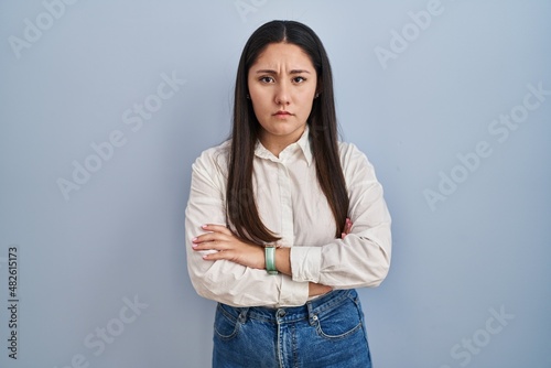 Young latin woman standing over blue background skeptic and nervous, disapproving expression on face with crossed arms. negative person.