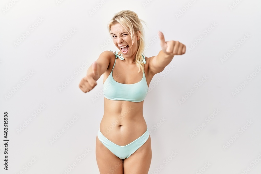 Young caucasian woman wearing bikini over isolated background approving doing positive gesture with hand, thumbs up smiling and happy for success. winner gesture. Young caucasian woman wearing bikini over isolated background approving doing positive gesture with hand, thumbs up smiling and happy for success. winner gesture.