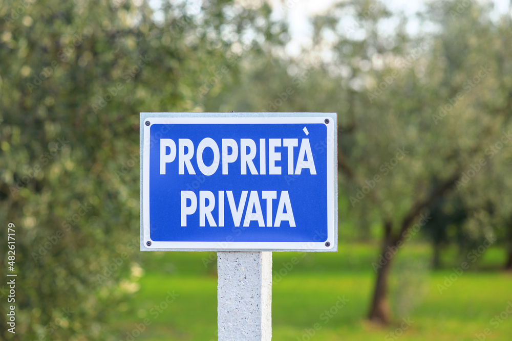 Blue sign with the inscription Private property in Italian outdoor in olive grove with olive trees blurred background Blue sign with the inscription Private property in Italian outdoor in olive grove with olive trees blurred background