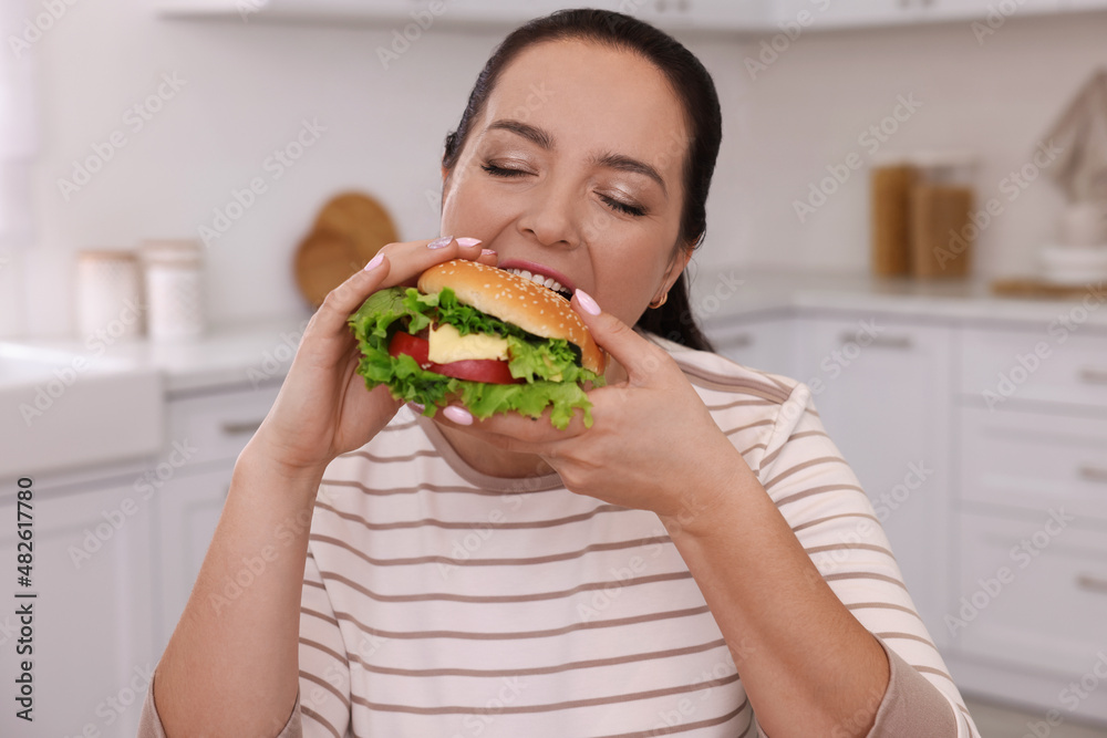 Overweight woman eating burger in kitchen at home Overweight woman eating burger in kitchen at home