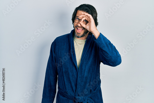 Middle age caucasian man wearing bathrobe and glasses doing ok gesture with hand smiling, eye looking through fingers with happy face.