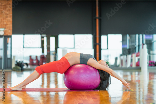 Portrait of young long dark-haired pregnant barefoot woman stretching on fit ball on mat. Doing yoga bandhasana pose in sport uniform. Light sunny day side view. Windows gym studio photo
