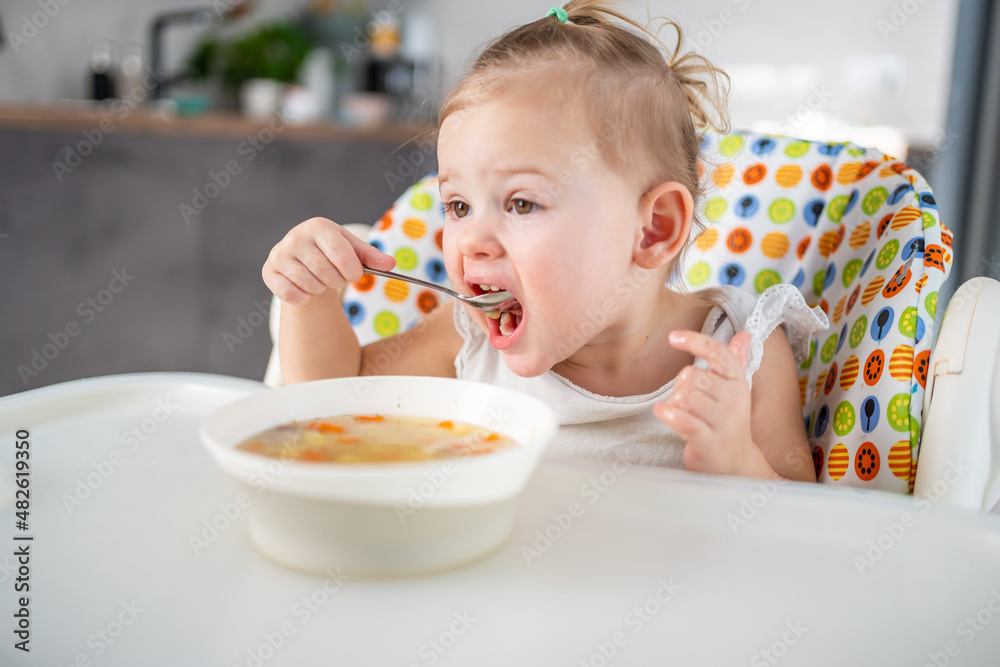 Cute baby girl toddler sitting in the high chair and eating her lunch soup at home kitchen. Cute baby girl toddler sitting in the high chair and eating her lunch soup at home kitchen.