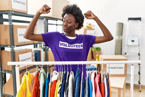 African young woman wearing volunteer t shirt at donations stand showing arms muscles smiling proud. fitness concept.