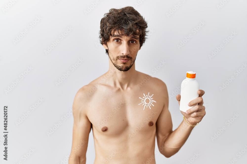 Fototapeta premium Young hispanic man standing shirtless holding sunscreen lotion relaxed with serious expression on face. simple and natural looking at the camera.