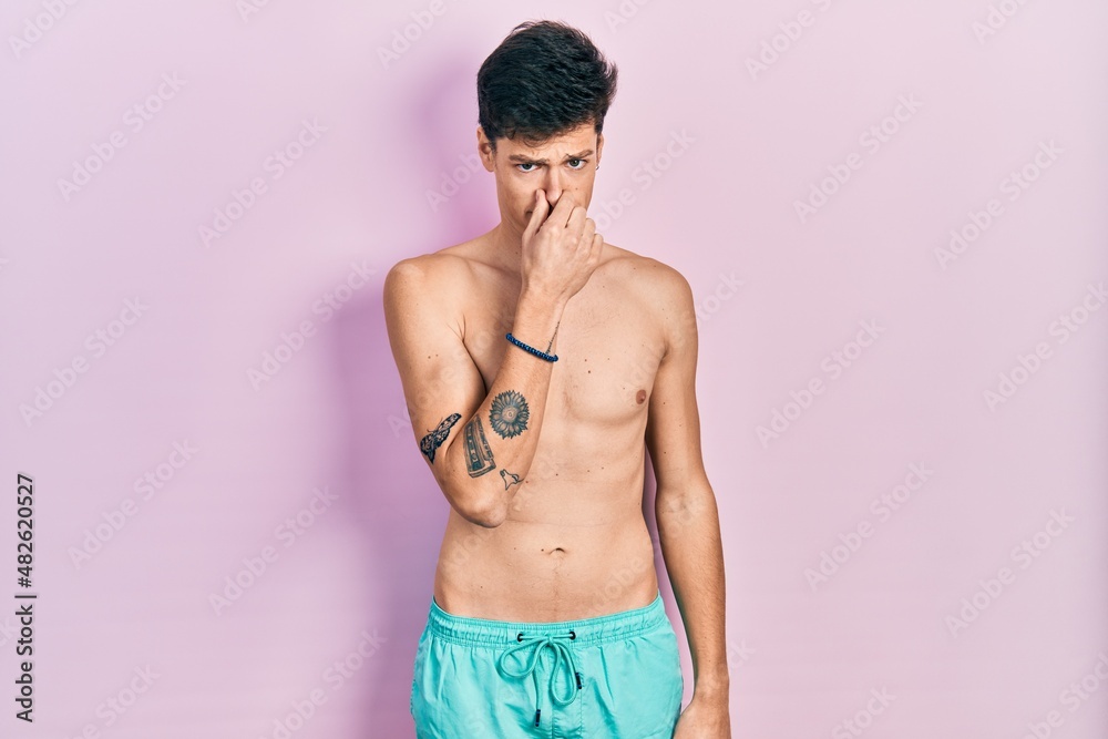 Young hispanic man wearing swimwear shirtless smelling something stinky and disgusting, intolerable smell, holding breath with fingers on nose. bad smell Young hispanic man wearing swimwear shirtless smelling something stinky and disgusting, intolerable smell, holding breath with fingers on nose. bad smell