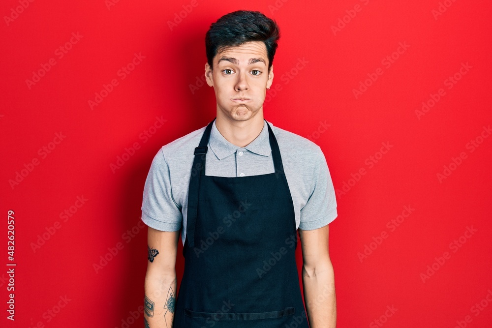 Young hispanic man wearing apron puffing cheeks with funny face. mouth inflated with air, crazy expression. Young hispanic man wearing apron puffing cheeks with funny face. mouth inflated with air, crazy expression.