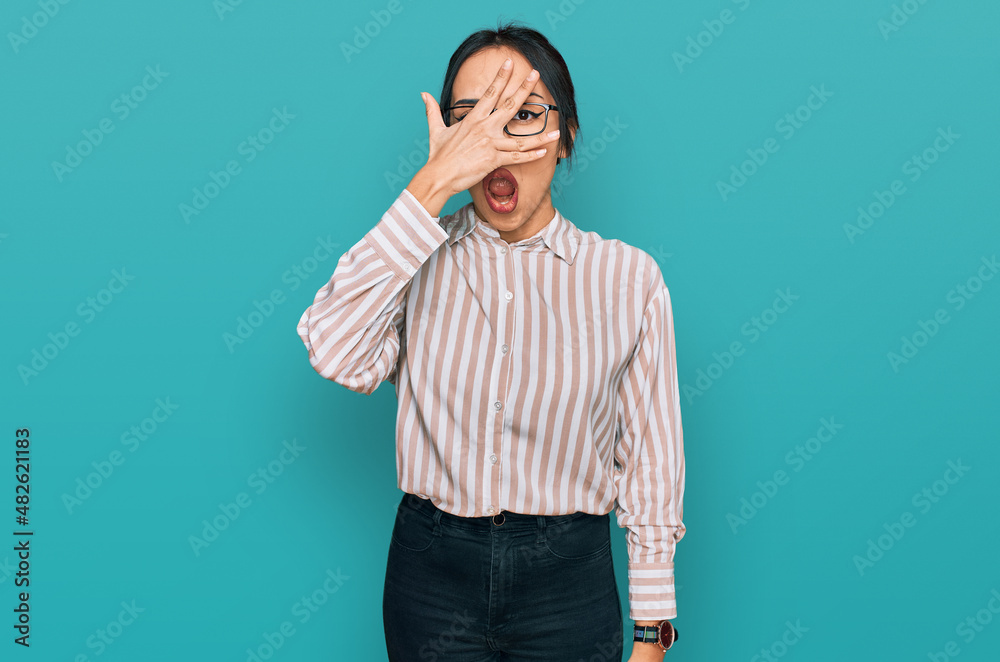 Young hispanic girl wearing casual clothes and glasses peeking in shock covering face and eyes with hand, looking through fingers with embarrassed expression. Young hispanic girl wearing casual clothes and glasses peeking in shock covering face and eyes with hand, looking through fingers with embarrassed expression.