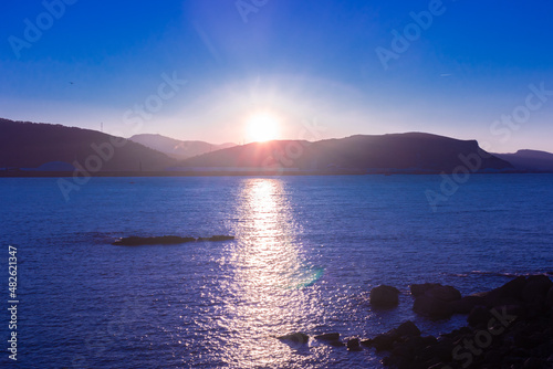 Vista de la silueta de montaña al horizonte sobre el mar al anochecer con cielo azulado y el sol reflejado en el mar creando un camino hasta nosotros