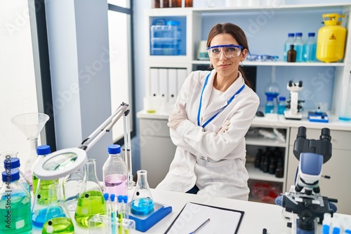 Young hispanic woman wearing scientist uniform standing with arms crossed gesture at laboratory