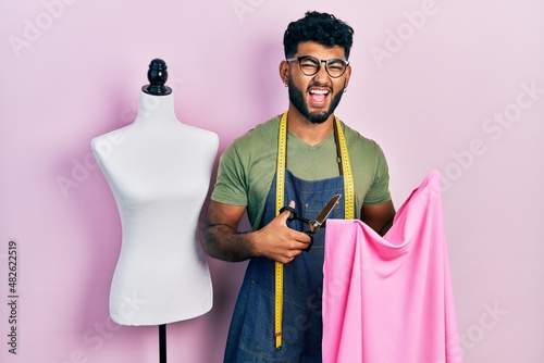 Arab man with beard dressmaker designer holding scissors and cloth smiling and laughing hard out loud because funny crazy joke.