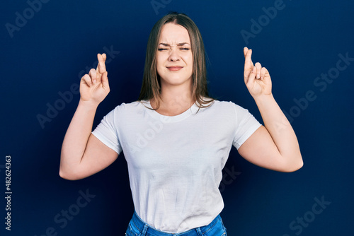 Young hispanic girl wearing casual white t shirt gesturing finger crossed smiling with hope and eyes closed. luck and superstitious concept.