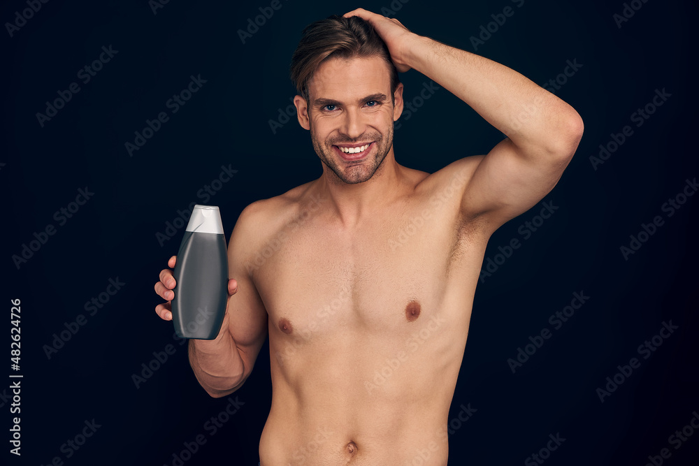 Handsome young man isolated. Portrait of shirtless muscular man is standing on dark blue background with shampoo in hands. Men care concept.
