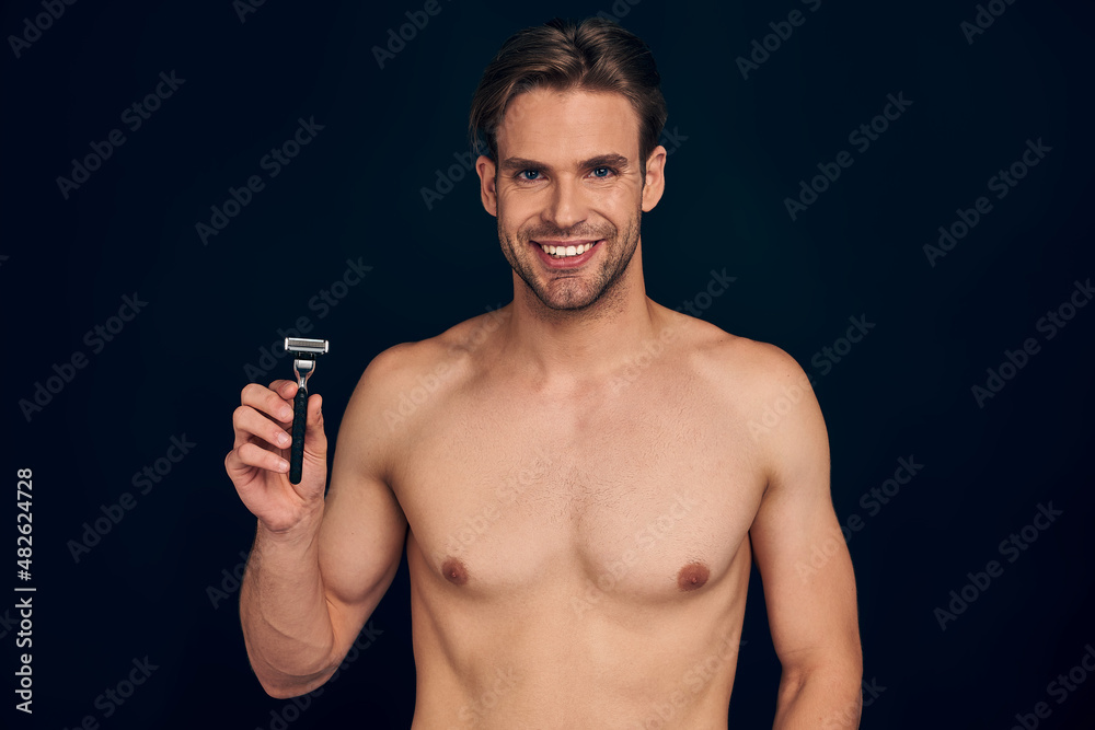 Handsome young bearded man isolated. Portrait of topless muscular man is standing on dark blue background with razor in hand . Men care concept