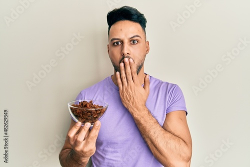 Young arab man holding raisins bowl covering mouth with hand, shocked and afraid for mistake. surprised expression