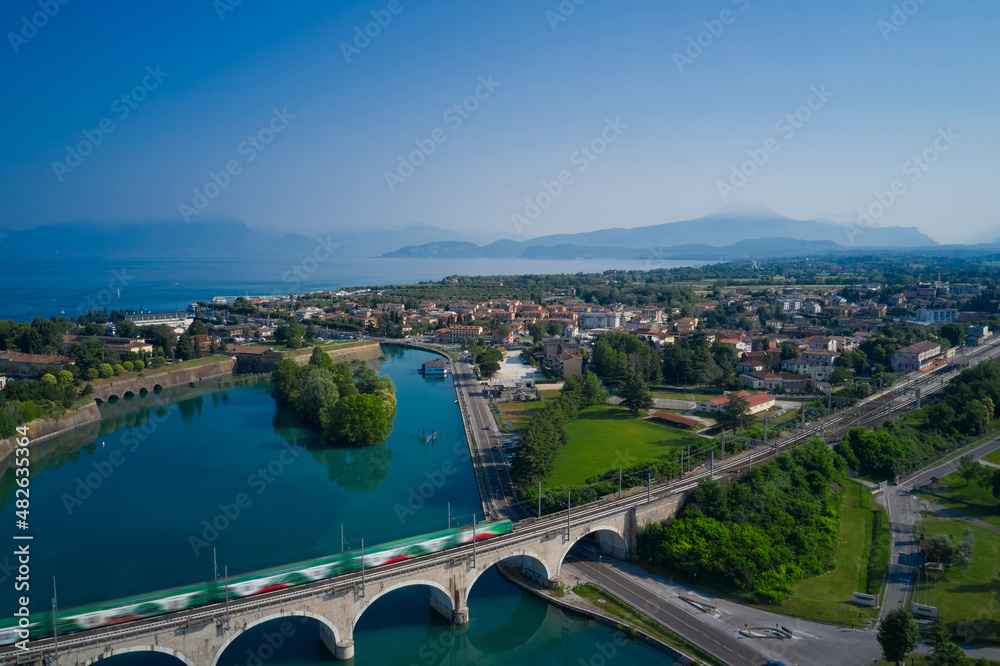 Peschiera del Garda, Italy. Train passes an arched railway bridge over a river in the background Lake Garda Italy.