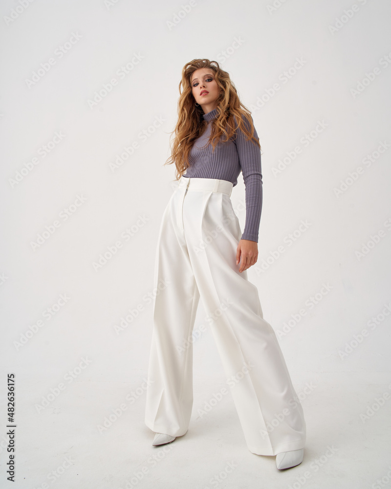 Pretty woman with stylish makeup and long flowing hair wearing sweater and loose trousers holds hand on hip on white background Pretty woman with stylish makeup and long flowing hair wearing sweater and loose trousers holds hand on hip on white background