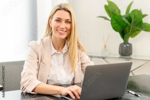 Smiling business woman using laptop for remote work from home. Confident lady checking emails, conducting business correspondence while sitting at the table, looks at the camera
