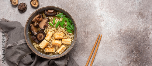 Asian vegan ramen noodle soup with roasted tofu cheese and shiitake mushrooms in a bowl on brown grunge background. Top view. Selective Focus. Banner.