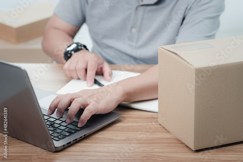 A man typing on laptop and check note list about the delivery products quickly and on time and there was a parcel box on the table to specify the address of the recipient