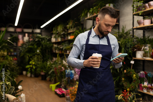an employee in a flower shop with a glass of coffee watches the news on his phone