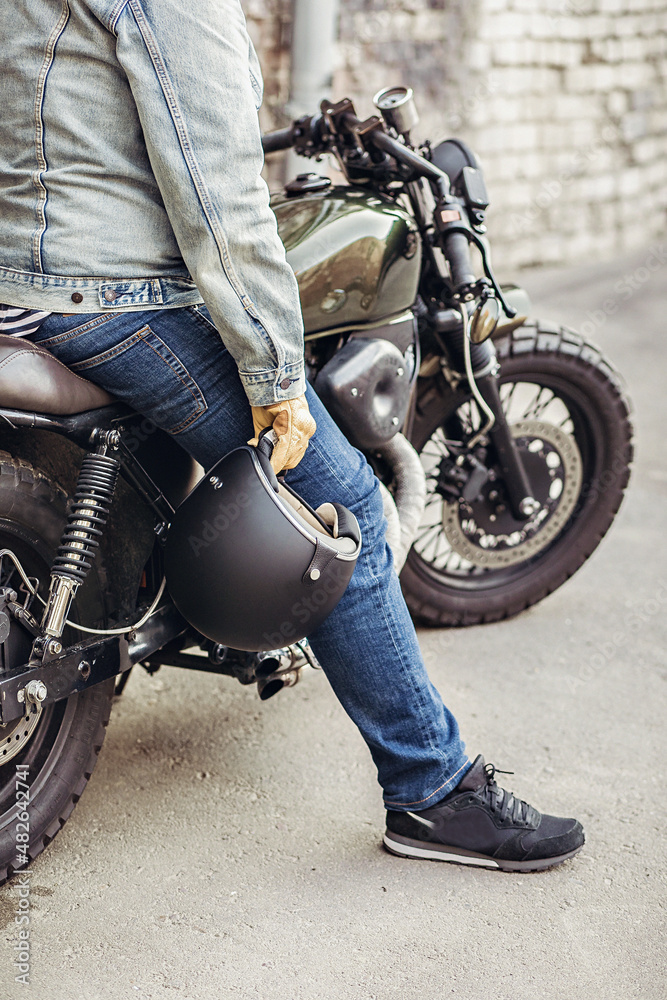 Close up attractive male model wearing denim clothes and gloves sitting on a bike and holding a helmet in his hands. Close up motorcycle detail. Concept of masculinity