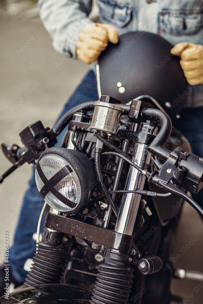 Fototapeta premium Extreme brutal motorcycle driver in denim clothes and gloves holding a black helmet and sitting on a motorcycle. Motorcycles to order. Speed concept.