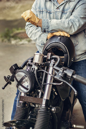 Master courageous motorcycle driver in denim clothes and gloves holding a black helmet and sitting on a motorcycle. Motorcycles to order. Sports concept.