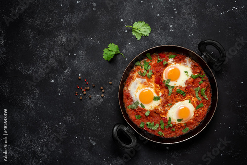 Shakshuka eggs in a pan with toast on a black concrete background. Poached eggs in a spicy tomato pepper sauce. Traditional Jewish scrambled eggs. Top view, flat lay.
