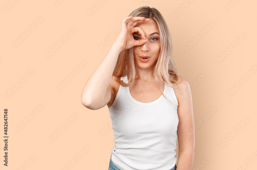 Joyful blonde caucasian female looking at camera through fingers in okay gesture isolated on color background. Joyful blonde caucasian female looking at camera through fingers in okay gesture isolated on color background.