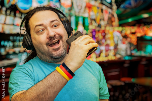 An unshaven middle-aged man in wireless headphones cheerfully sings into a mobile phone in a bar after work. Bracelet in the colors of the German flag.