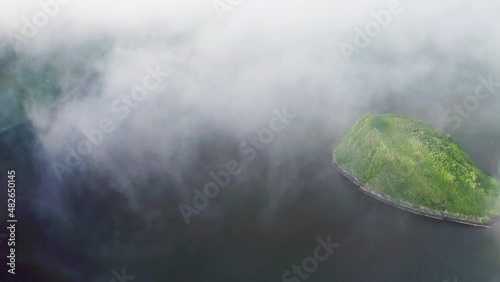 Aerial panorama. Green islet hide by dense mist clouds flying above dark water 