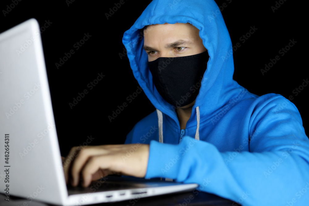 Man in mask and blue hoodie sitting with laptop on black background. Concept of cyber crime, hacking and safety work during coronavirus Man in mask and blue hoodie sitting with laptop on black background. Concept of cyber crime, hacking and safety work during coronavirus