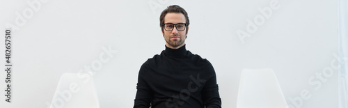 young man in eyeglasses and black uniform looking at camera near chairs on grey background, banner. © LIGHTFIELD STUDIOS
