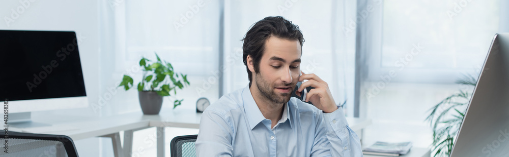 positive businessman talking on mobile phone near computer monitors in office, banner. positive businessman talking on mobile phone near computer monitors in office, banner.