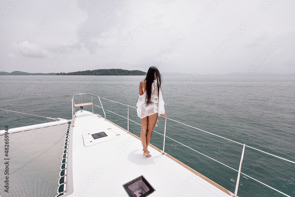 Pretty turned back lady in white clothes is looking at the water from a height, standing on her yacht, wide angle