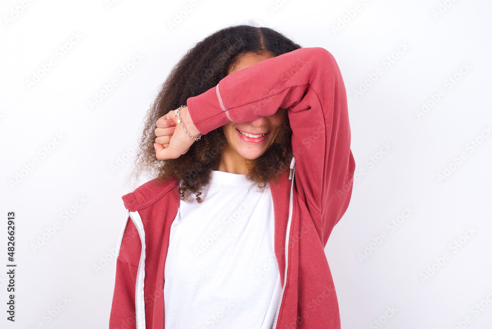 teenager girl wearing pink jacket standing against wite background covering eyes with arm smiling cheerful and funny. Blind concept.