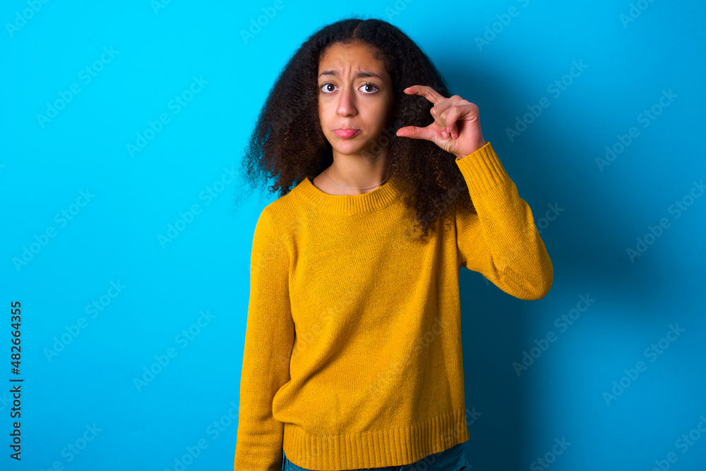beautiful teenager girl wearing yellow sweater standing against blue background purses lip and gestures with hand, shows something very little. beautiful teenager girl wearing yellow sweater standing against blue background purses lip and gestures with hand, shows something very little.